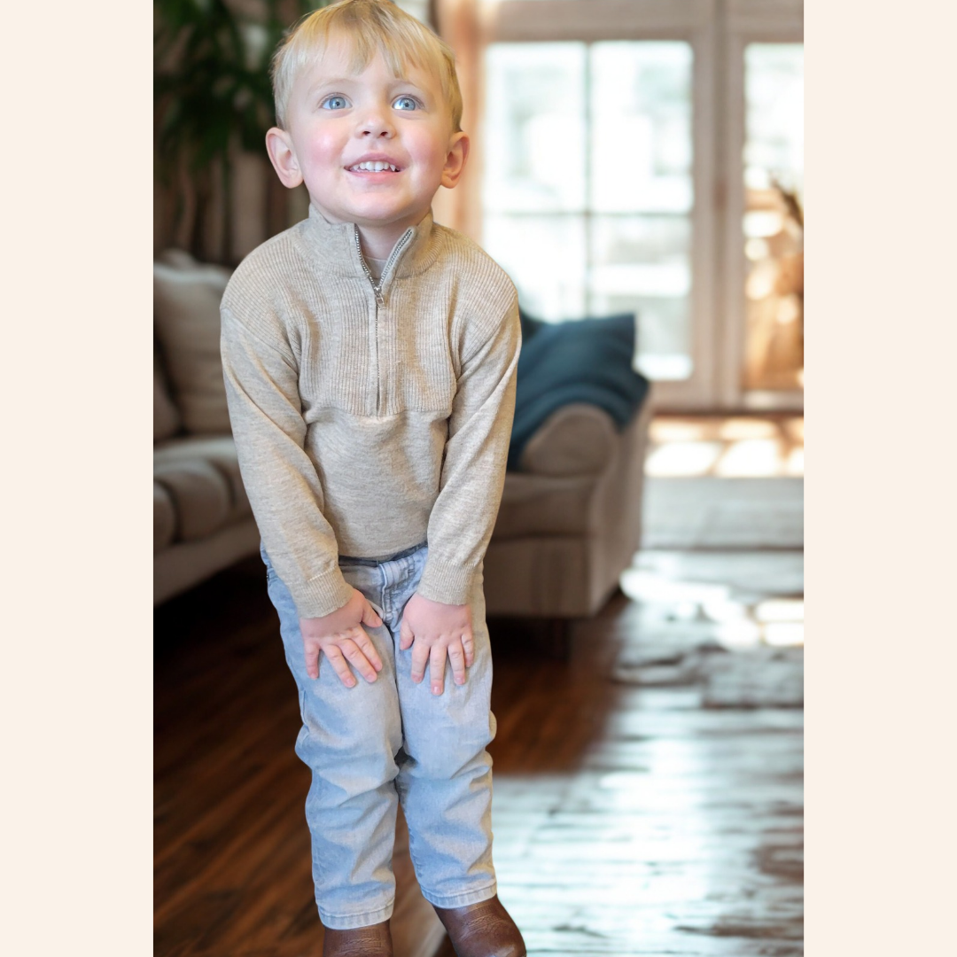 Child wearing a beige sweater and light blue jeans standing in a living room.