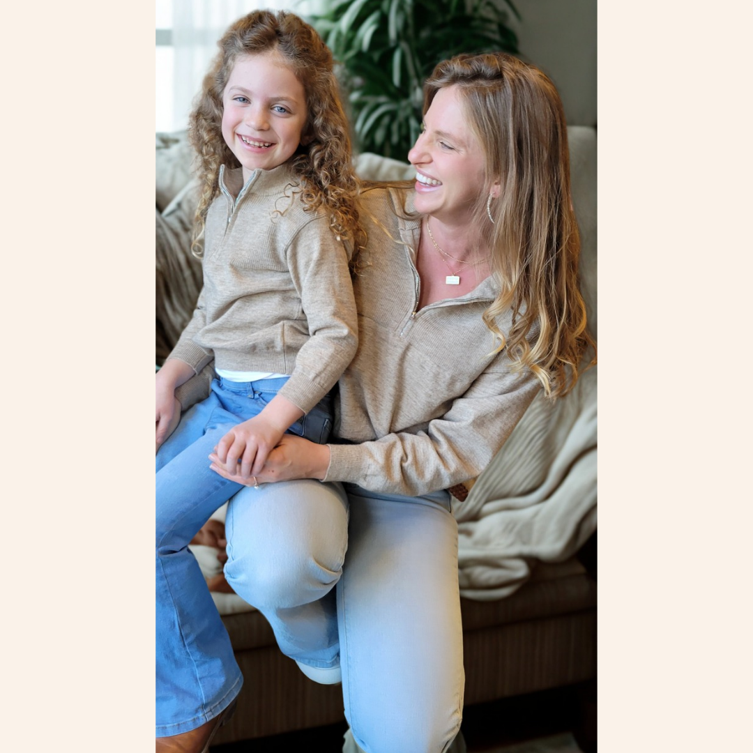 Woman and young girl sitting together on a couch, both wearing beige sweaters.