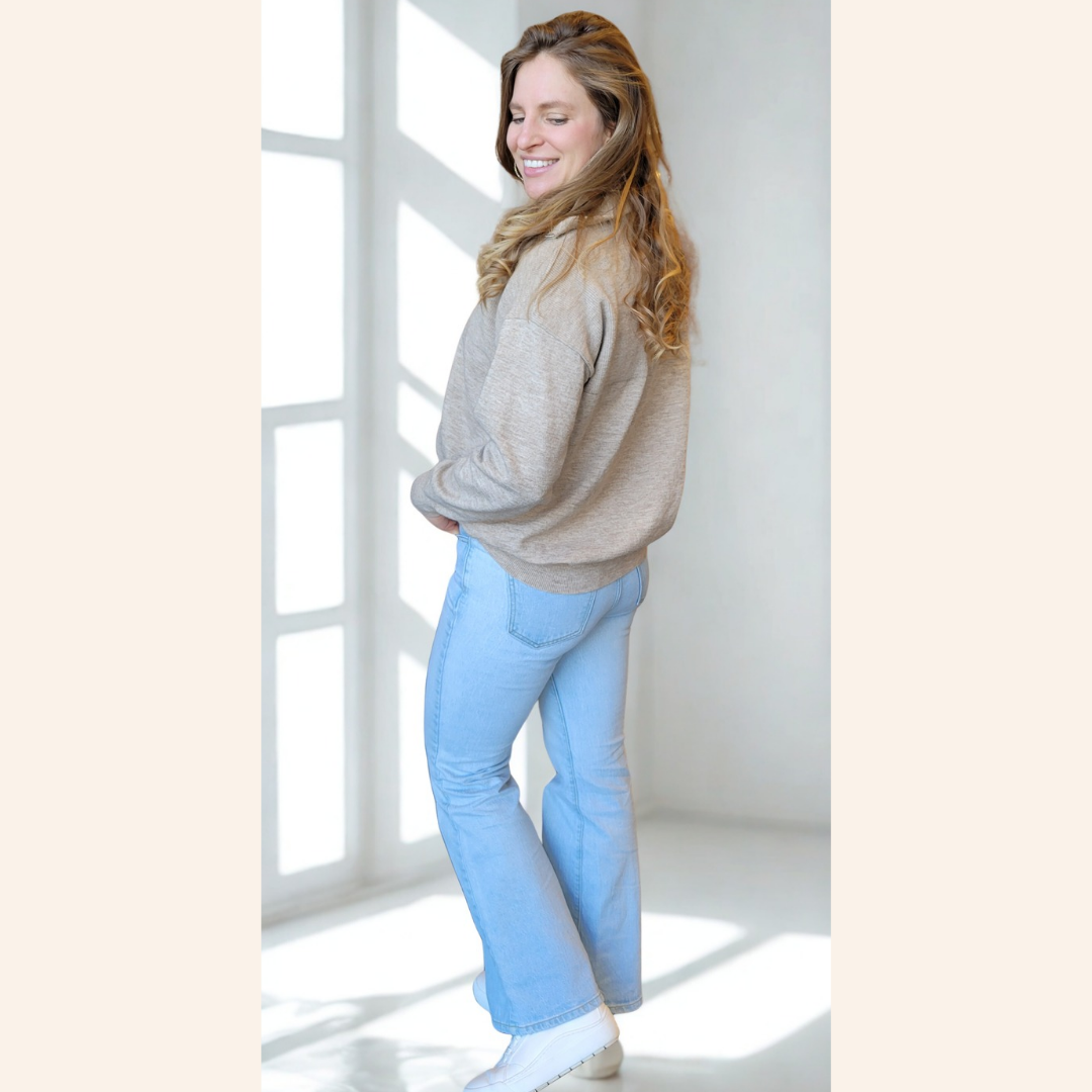 Woman wearing a beige sweater and light blue pants standing in a bright room with large windows.