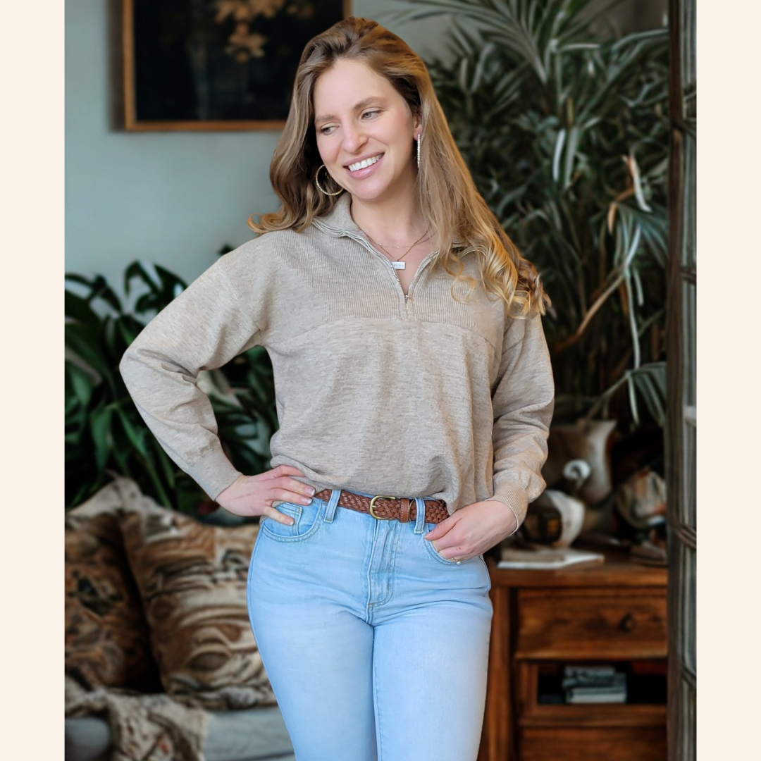 Woman wearing a beige sweater and light blue jeans indoors with plants and furniture in the background