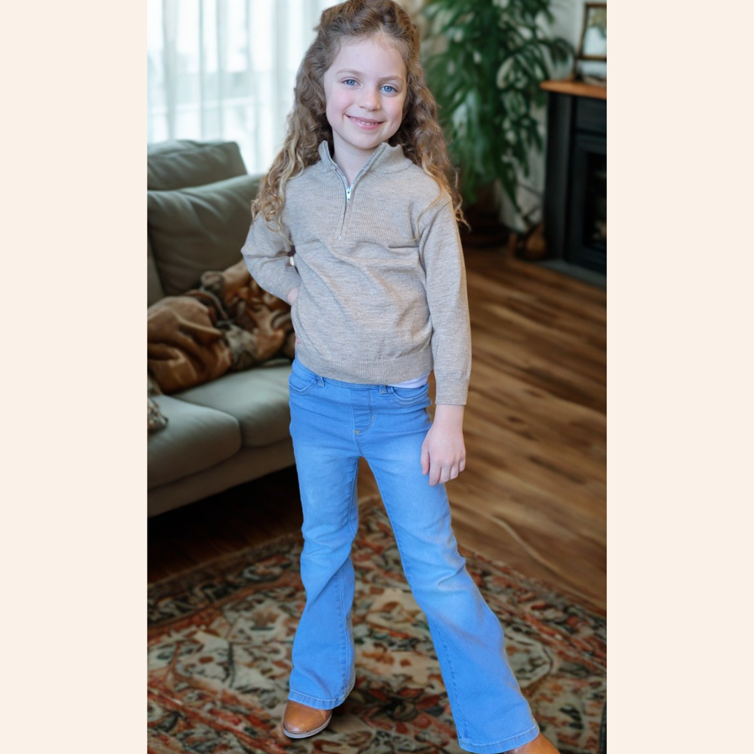Young girl wearing a beige sweater and blue jeans standing in a living room.