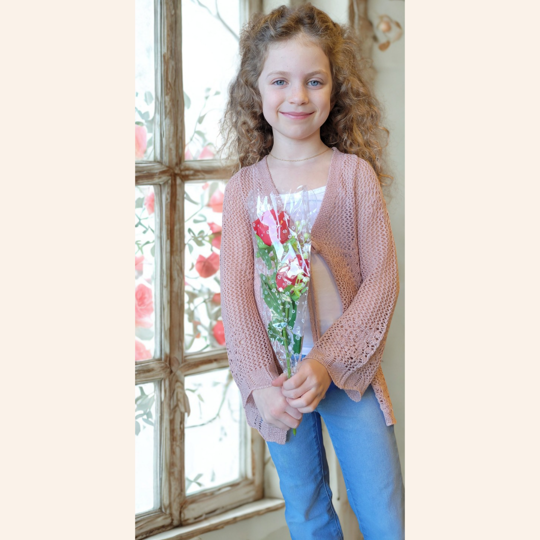 Young girl wearing a pink cardigan over a floral shirt, standing in front of a window with floral decorations.