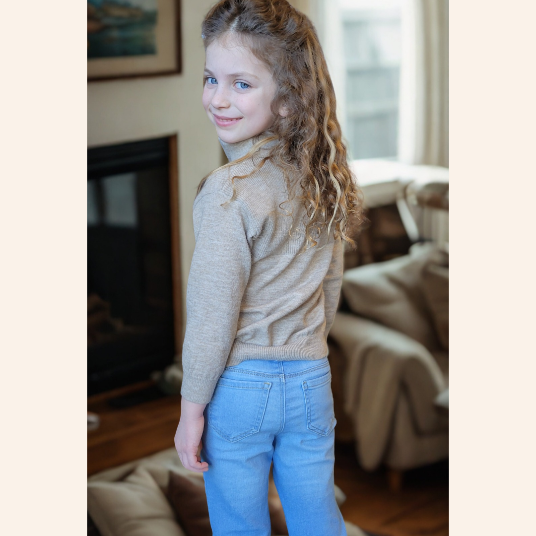 Young girl wearing a beige sweater and blue jeans standing in a living room.