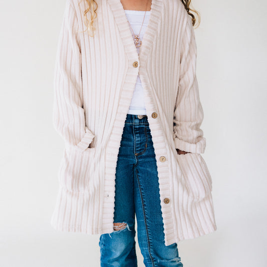 Child wearing a long beige cardigan over a white shirt and blue jeans on a plain background