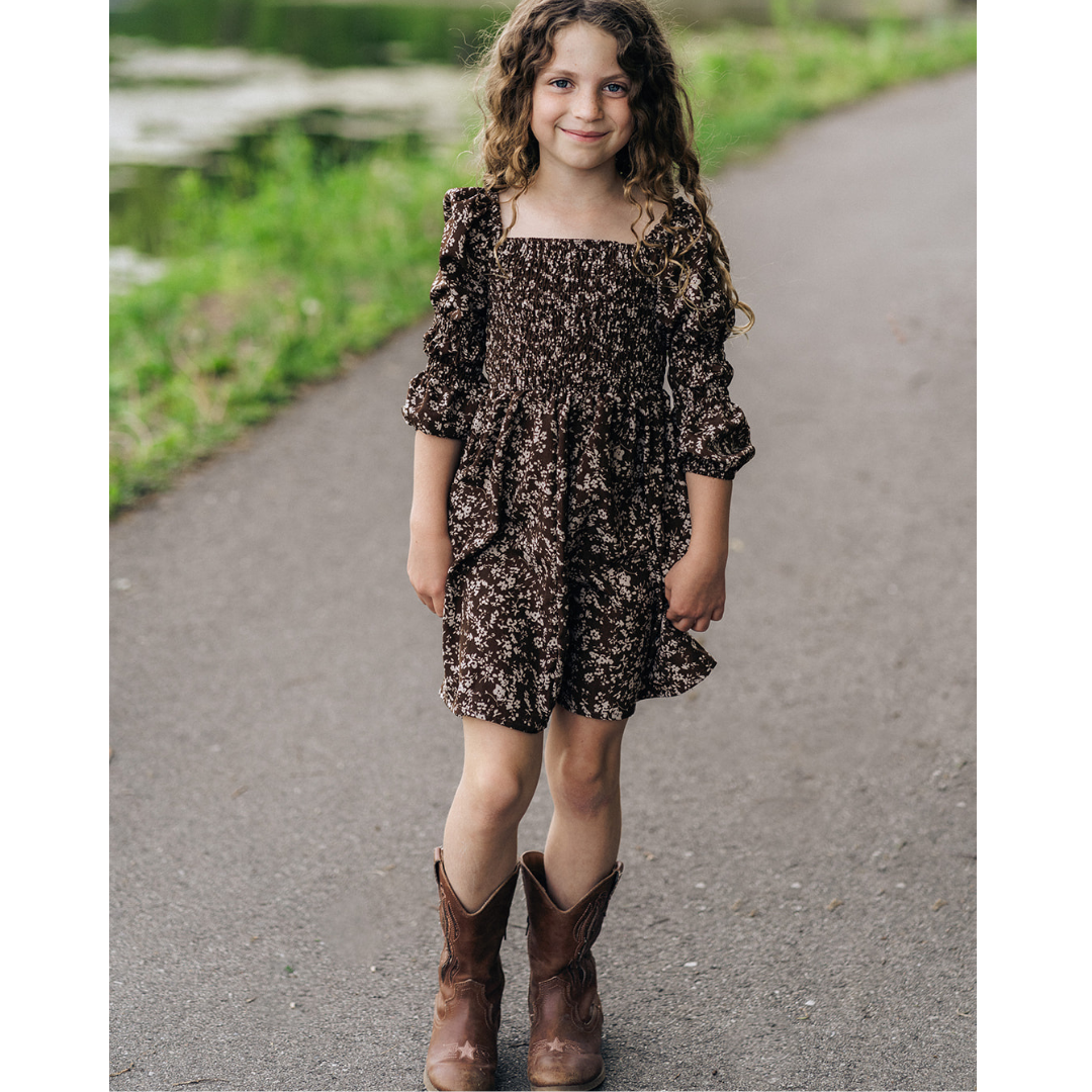 Young girl in a floral dress and brown boots standing on a path with greenery.