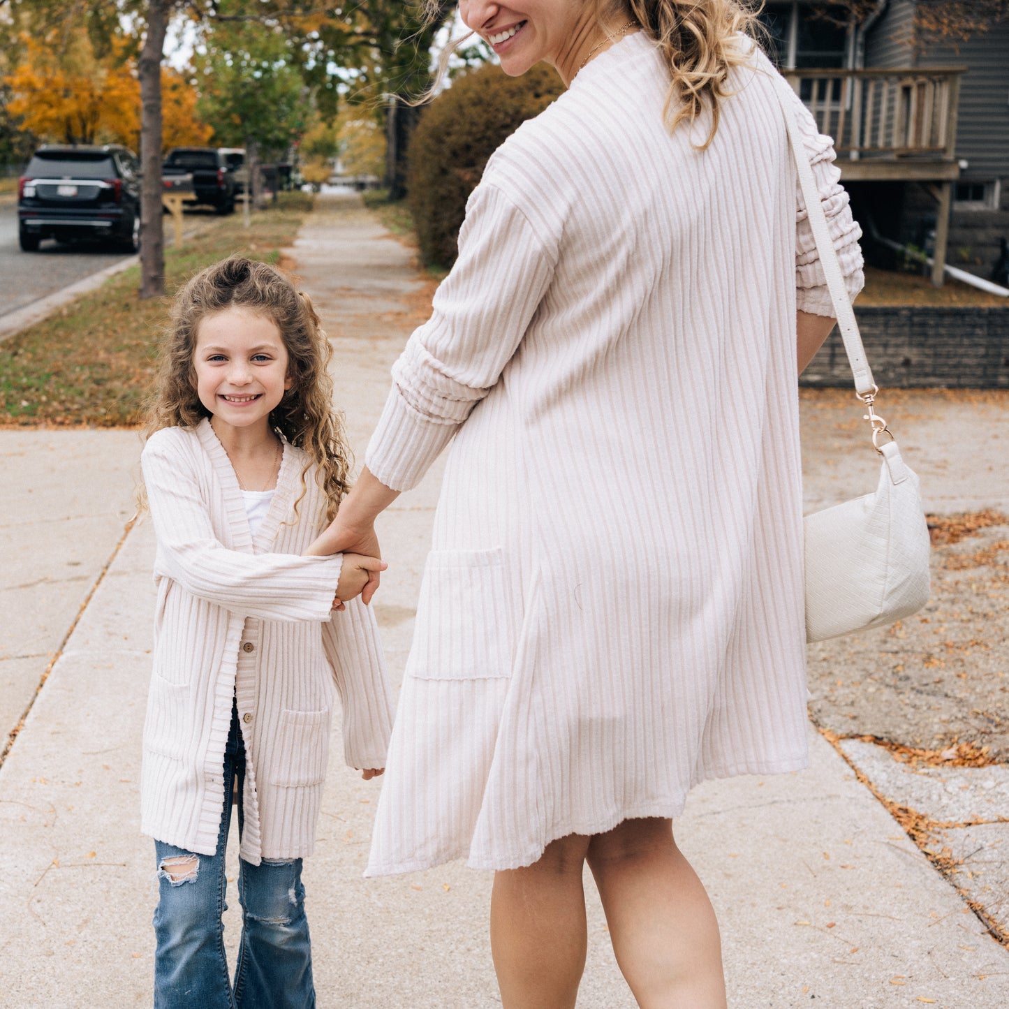 Woman and child holding hands on a sidewalk with autumn leaves and houses in the background